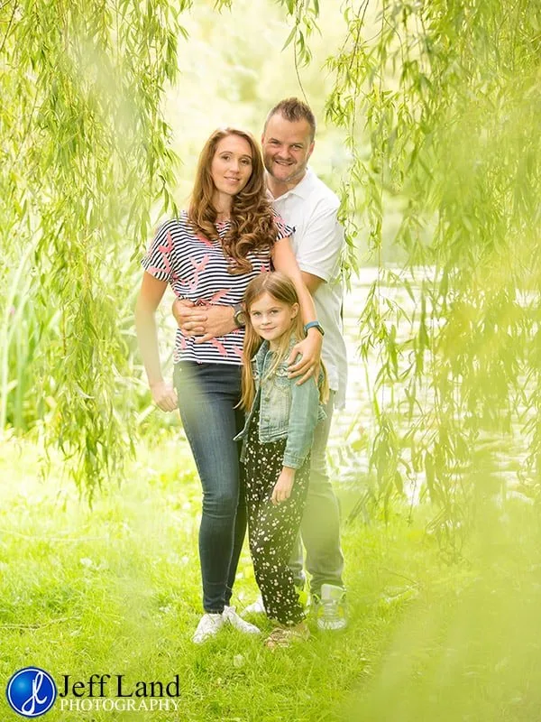 A happy family of three posing together outdoors under a tree.