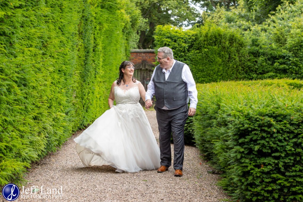 A bride and groom walk hand in hand down a gravel pathway surrounded by tall, green hedges, creating a picturesque moment worthy of glowing testimonials.