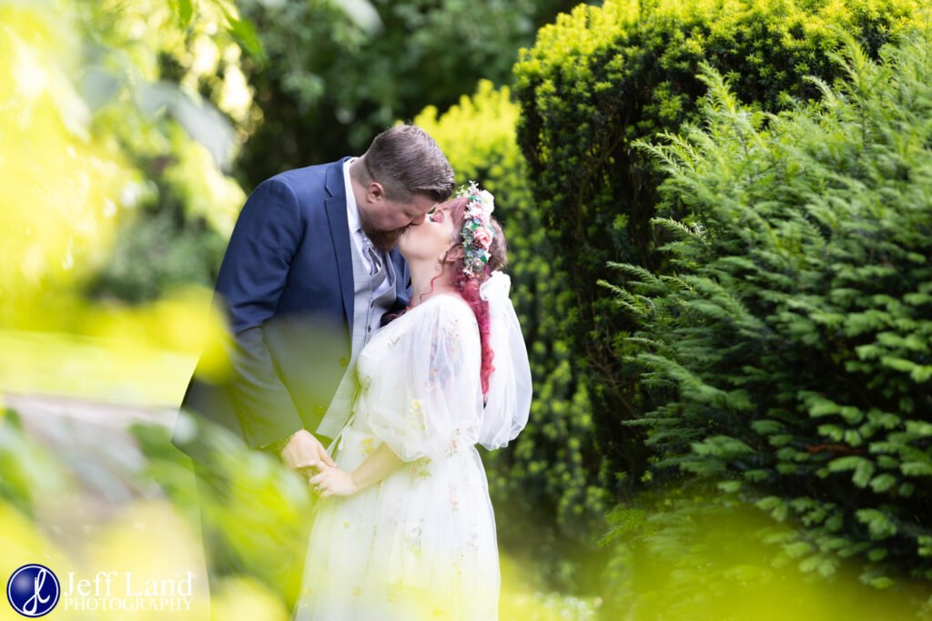 A couple kisses in a lush garden, surrounded by greenery. The woman wears a floral crown and white dress, while the man is in a dark jacket. Photo by Jeff Land Photography, known for capturing memorable moments that clients rave about in their testimonials.